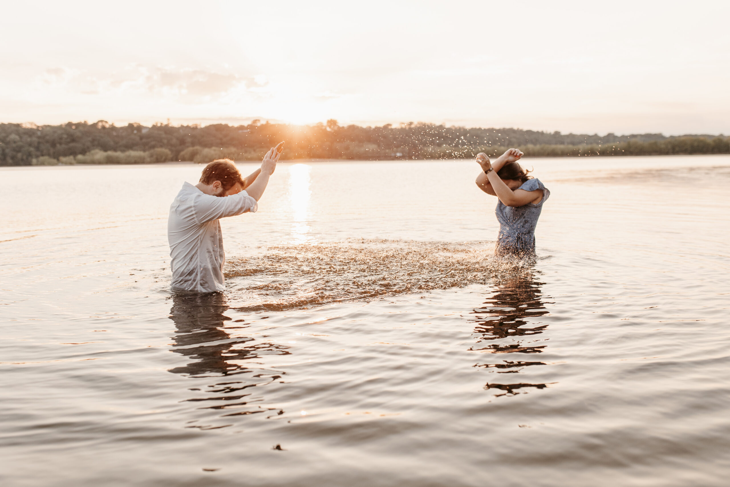 man and woman splashing each other in a lake fully clothed at sunset for their anniversary photos
